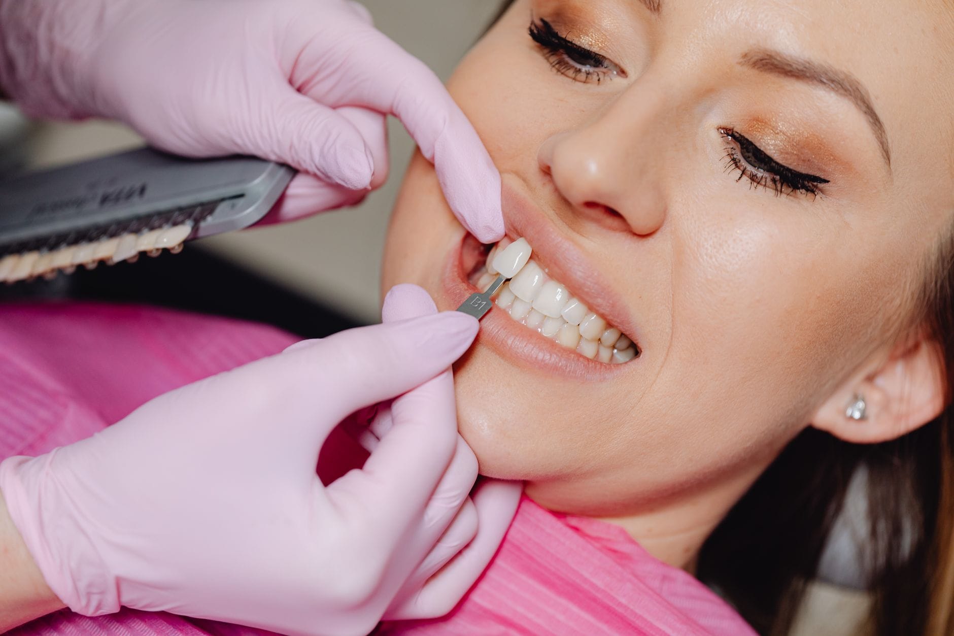 Dental professional in pink gloves compares tooth color using a shade guide against a smiling patient's teeth.