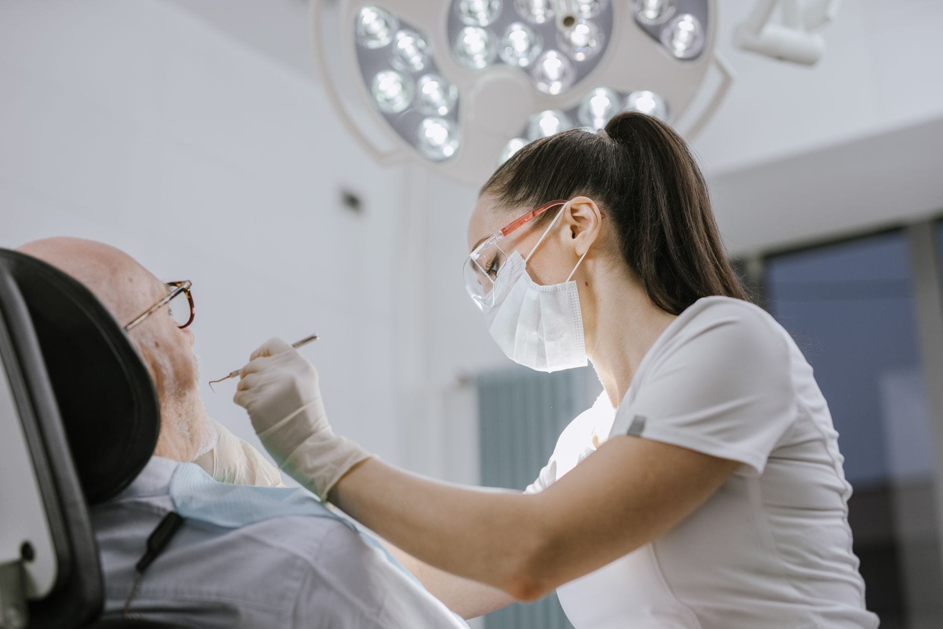 Dental professional in PPE treats an elderly patient in a reclining chair under bright surgical lights. (informative: dental procedure)