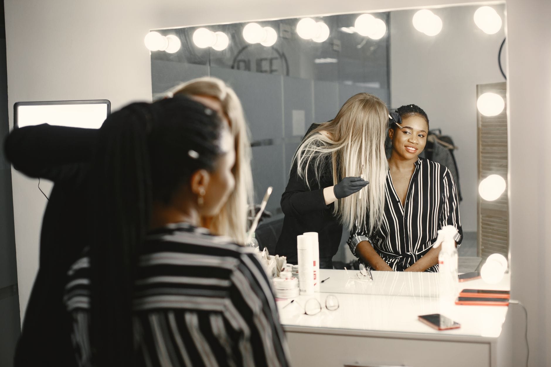 Two women at a makeup vanity with a large illuminated mirror, one with blonde hair helping the other in a striped shirt smile at the reflection